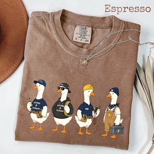 May include: Espresso-colored t-shirt featuring a cartoon design of four geese dressed as electricians, each with tools and different hats. The text "Salty Goose Electrician" is on the shirts. The shirt has a crew neck and short sleeves.