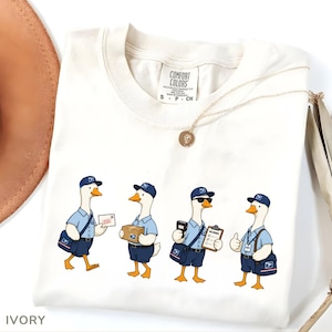 Puede incluir: Camiseta Comfort Colors de color marfil con un dise&ntilde;o de dibujos animados de cuatro gansos vestidos de carteros. Los gansos se muestran en varias poses, llevando correo, paquetes y portapapeles. Tambi&eacute;n se ve un collar dorado y un sombrero marr&oacute;n.