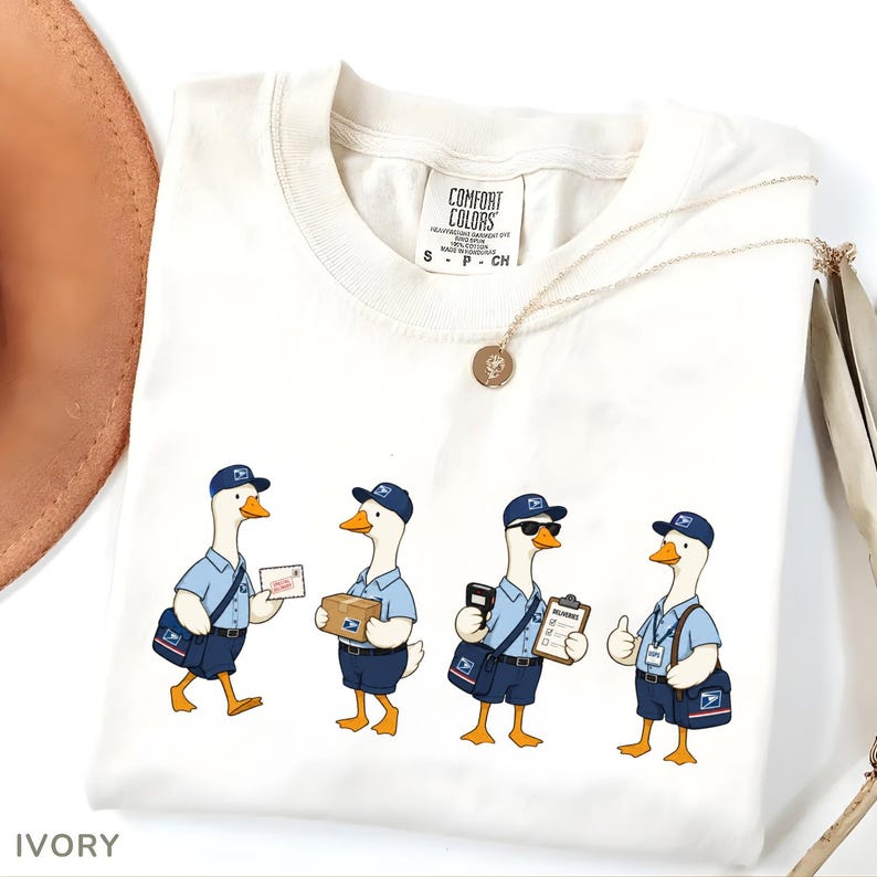 Puede incluir: Camiseta Comfort Colors de color marfil con un dise&ntilde;o de dibujos animados de cuatro gansos vestidos de carteros. Los gansos se muestran en varias poses, llevando correo, paquetes y portapapeles. Tambi&eacute;n se ve un collar dorado y un sombrero marr&oacute;n.