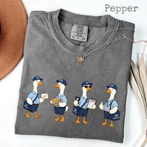 Puede incluir: Camiseta gris color "Pepper" con un dise&ntilde;o de dibujos animados de cuatro gansos vestidos de carteros. Cada ganso lleva uniforme azul, gorra y bandolera, y sostiene correo y paquetes.