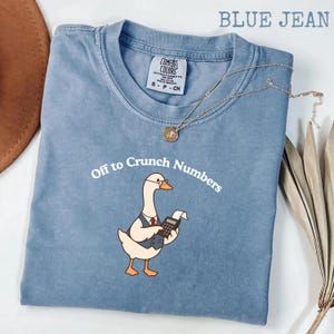 May include: A blue jean coloured t-shirt featuring a graphic of a white goose wearing a vest and glasses while holding a calculator. The text above the goose reads Off to Crunch Numbers in white font. The shirt has a Comfort Colors brand label.