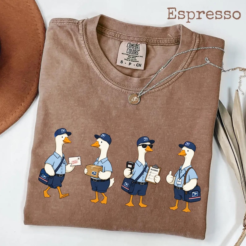 Puede incluir: Camiseta color espresso con un dise&ntilde;o divertido de cuatro gansos de dibujos animados vestidos de carteros. Cada ganso lleva un uniforme azul, llevando correo, paquetes y portapapeles. La camiseta tiene cuello redondo y mangas cortas.