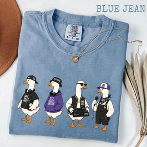 May include: A blue jean coloured t-shirt featuring a graphic of four geese styled as DJs. Each goose is dressed in various outfits, including hats, headphones, and sunglasses. The text "DJ SILLY GEESE" is printed on the shirt. A gold necklace is draped across the shirt.