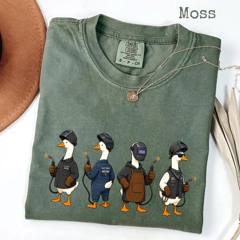 May include: A moss green crew neck t-shirt featuring a graphic of four white geese dressed as welders, each wearing a welding helmet and holding a welding torch. The shirt has a Comfort Colors label at the inner neckline and a gold-toned necklace.