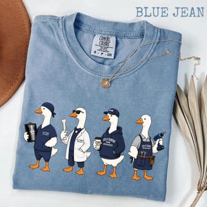 May include: Blue jean colored t-shirt with a graphic of four cartoon geese dressed in various orthopedics-related outfits. The text "Silly Goose Orthopedics" is on the shirt. A gold necklace with a pendant is draped across the shirt.