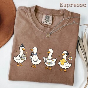 Peut inclure: T-shirt couleur expresso avec une rangée de canards de dessin animé, chacun avec un accessoire unique : un chapeau, une loupe, un appareil photo et un casque. Le t-shirt a une étiquette Comfort Colors et un collier argenté.