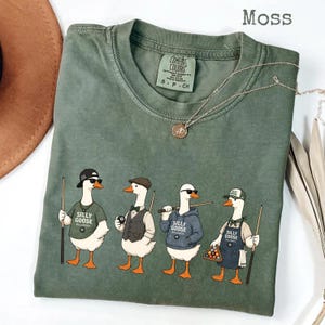Peut inclure: T-shirt vert mousse avec un motif de quatre oies habillées en joueurs de billard, chacune avec une queue de billard. Le texte "Silly Goose Billiards" est sur les chemises. Le t-shirt a un col rond et des manches courtes. Un collier argenté est drapé sur le t-shirt.