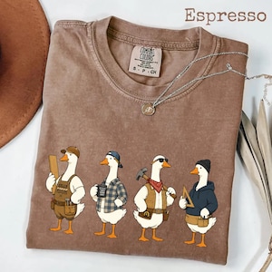 May include: An espresso-coloured short-sleeve t-shirt featuring a graphic of four cartoon geese dressed as construction workers. The geese are wearing hats, vests, and holding tools like a wooden board, a mug, a hammer, and a triangle ruler.
