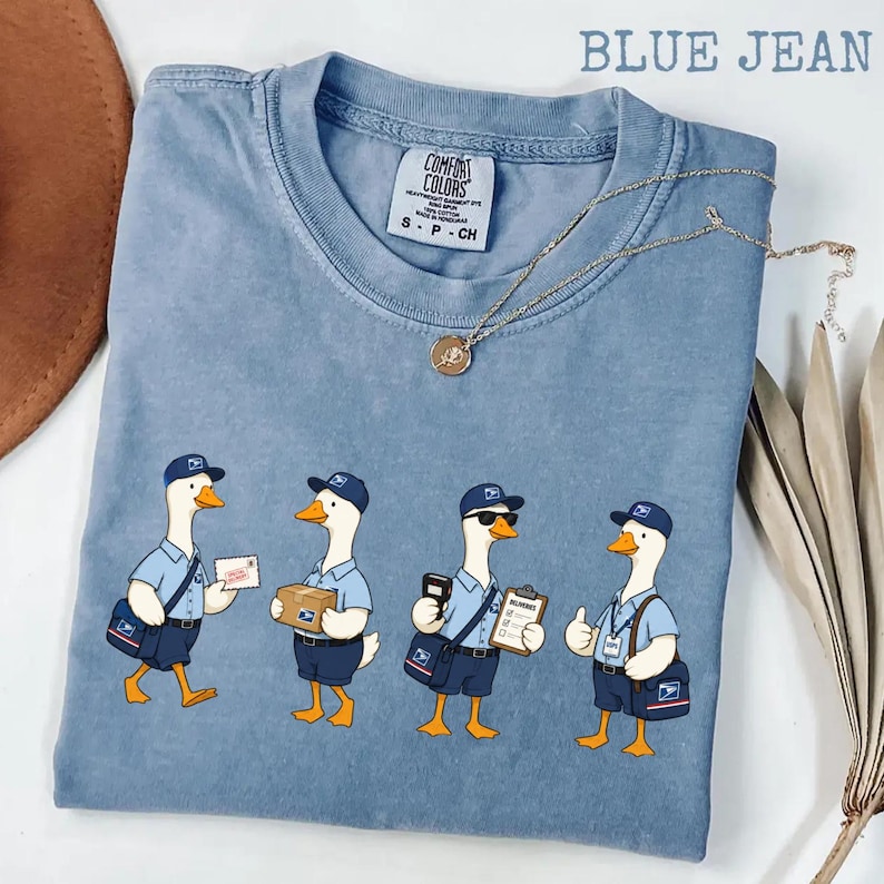 Puede incluir: Camiseta azul vaquero con un gr&aacute;fico de cuatro gansos de dibujos animados vestidos como carteros. Cada ganso lleva un uniforme azul, sombrero y bandolera. Los gansos sostienen correo, un paquete, una carpeta y hacen una se&ntilde;al de aprobaci&oacute;n. Un collar dorado est&aacute; sobre la camisa.