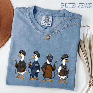 May include: A blue jean colored t-shirt with a graphic print of four white geese wearing welding helmets and protective gear, each holding a welding torch. The shirt features a Comfort Colors brand label at the neck and a crew neckline.