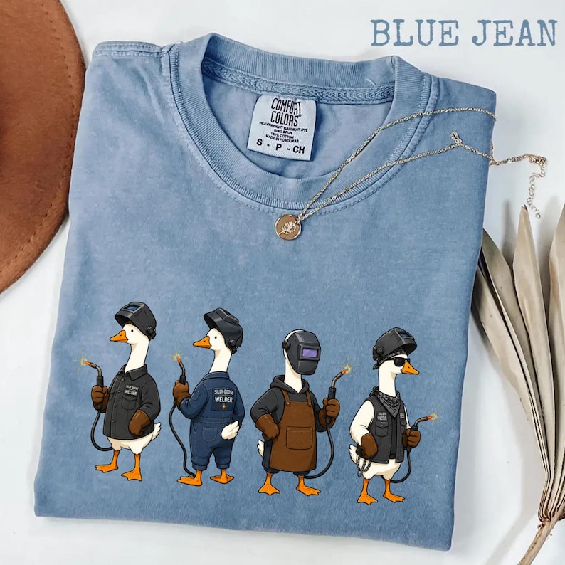 May include: A blue jean colored t-shirt with a graphic print of four white geese wearing welding helmets and protective gear, each holding a welding torch. The shirt features a Comfort Colors brand label at the neck and a crew neckline.