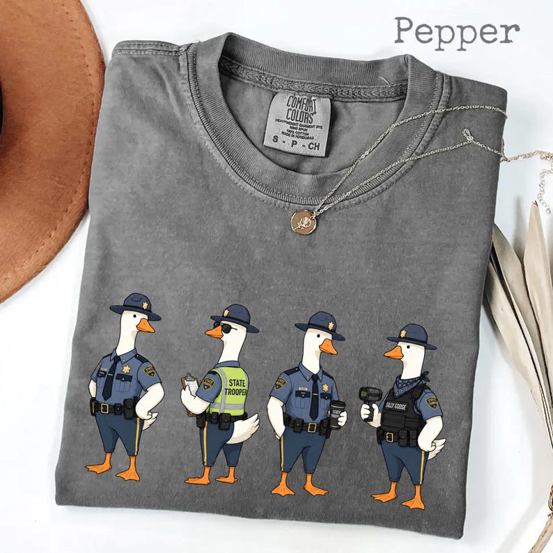 May include: A pepper-colored t-shirt featuring a graphic of four geese dressed as state troopers. The geese wear blue uniforms, hats, and tactical gear. One goose wears sunglasses and a high-visibility vest. The shirt has a Comfort Colors label.