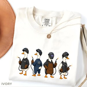 May include: An ivory t-shirt featuring a graphic of four white geese dressed as welders. Each goose wears a welding helmet and holds a welding torch. One goose wears a black jacket, one a blue jumpsuit, one a brown apron, and one a black vest.