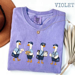 Puede incluir: Camiseta violeta Comfort Colors con un dise&ntilde;o caprichoso de cuatro gansos carteros. Cada ganso viste un uniforme postal, llevando correo, paquetes y portapapeles. Tambi&eacute;n se ven un collar dorado y un sombrero marr&oacute;n.