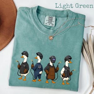 May include: A light green t-shirt featuring a graphic of four geese dressed as welders, each wearing a welding helmet and holding a welding torch. The shirt has a crew neckline and a Comfort Colors brand label on the inside of the collar.