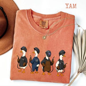 May include: A terracotta-colored crew neck t-shirt featuring a graphic of four cartoon ducks dressed as welders, each wearing a welding helmet and holding a torch. The shirt has a Comfort Colors label at the inner neckline and the text YAM above it.