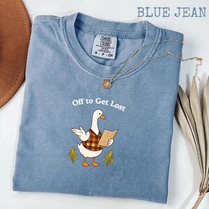 May include: A blue jean colored t-shirt featuring a graphic of a white goose wearing a brown and orange plaid vest, holding an open book, with the text "Off to Get Lost" above it. The shirt has a crew neckline and a Comfort Colors brand label.