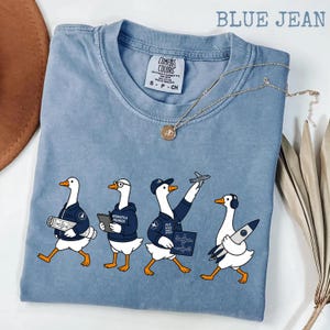 May include: Pale blue t-shirt featuring a cartoon design of four geese in navy blue jackets, each representing an aeronautical engineer. The geese are holding blueprints, tablets, and a rocket. A gold necklace with a pendant is draped across the shirt.