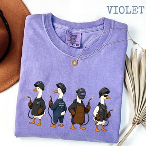 May include: A violet short-sleeved t-shirt featuring a graphic of four white geese wearing welding helmets and protective gear, each holding a welding torch. The shirt has a crew neckline and a Comfort Colors brand label on the inner collar.
