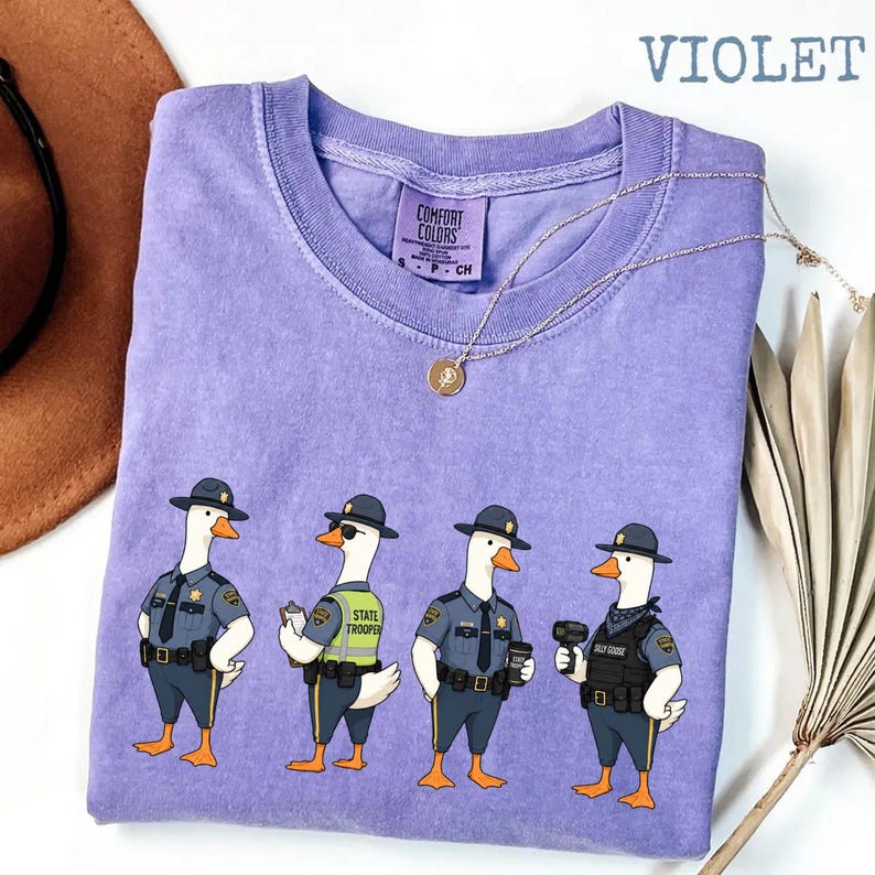 May include: A violet crew neck t-shirt featuring a graphic of four geese dressed as state troopers. The shirt has a Comfort Colors label at the inner neckline. The text VIOLET is printed in the upper right corner of the image.
