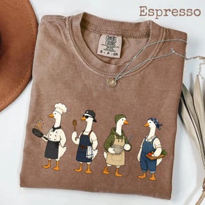 Peut inclure: T-shirt couleur expresso avec un motif de quatre oies vêtues en chefs, chacune tenant un ustensile de cuisine différent. Le t-shirt a un col rond et une étiquette Comfort Colors. Un collier argenté avec un pendentif est drapé sur le t-shirt.
