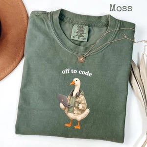 May include: A moss green crew neck t-shirt featuring a graphic of a white duck wearing a plaid shirt, green hoodie, and headphones while holding a laptop and a mug. The text "off to code" is printed above the duck in white, lowercase letters.