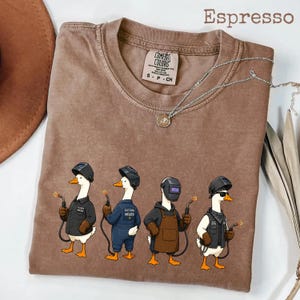 May include: An espresso-colored t-shirt featuring a graphic of four geese wearing welding helmets and protective gear, each holding a welding torch. The shirt has a crew neckline and a Comfort Colors brand label printed on the inside collar area.