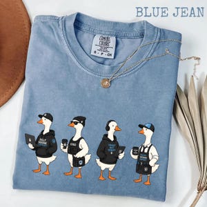 Peut inclure: Un t-shirt à manches courtes bleu jean avec un motif représentant quatre oies habillées en professionnels de la cybersécurité. Les oies portent des sweats à capuche, des chapeaux et des accessoires, tenant des ordinateurs portables et des tasses avec les textes Silly Goose, CISO, Trust But Verify et Patch Defend Repeat.