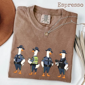 May include: An espresso-colored short-sleeve t-shirt featuring a graphic of four geese dressed as police officers. The geese wear blue uniforms, hats, and tactical gear. One goose wears sunglasses and a neon vest. The shirt has a Comfort Colors label.