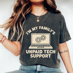 May include: Dark gray t-shirt with the text "I'M MY FAMILY'S UNPAID TECH SUPPORT" above a laptop graphic with gears on the screen. The shirt is a casual style.