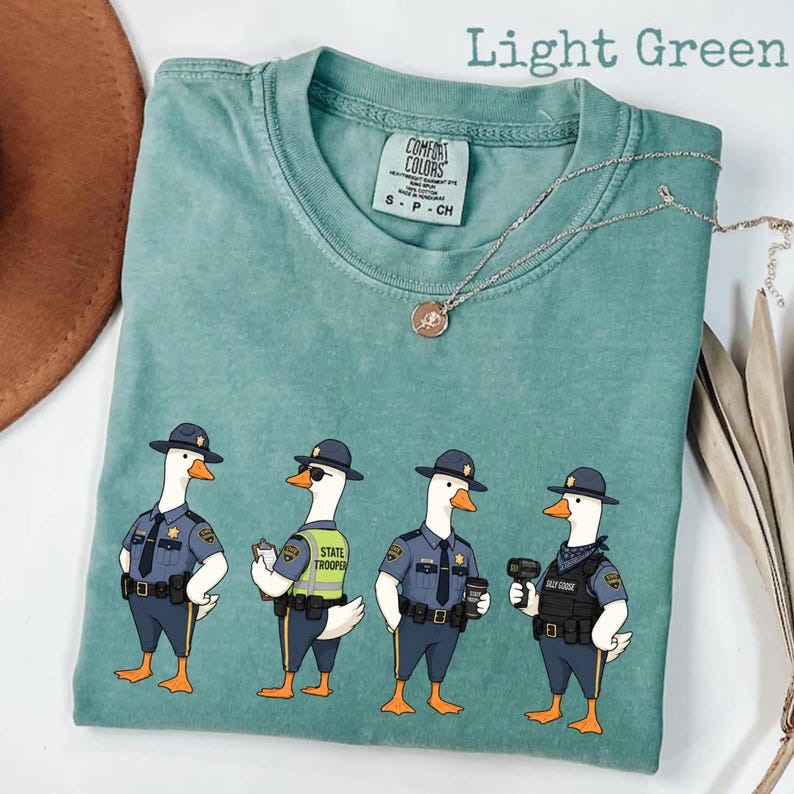 May include: A light green t-shirt featuring a graphic of four geese dressed as state troopers. The geese wear blue uniforms, hats, and various police gear including a vest, clipboard, and radio. The shirt has a Comfort Colors brand label.