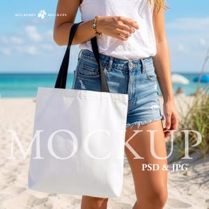 Puede incluir: Una bolsa de mano blanca con correas negras es sostenida por una persona en una playa. La bolsa tiene la palabra "MOCKUP" impresa en letras blancas grandes. El fondo incluye el océano y la arena.
