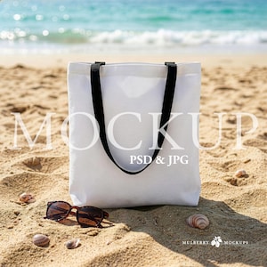 Puede incluir: Un bolso tote blanco con asas negras descansa sobre una playa arenosa, con gafas de sol y conchas cerca. El océano es visible en el fondo. La bolsa es una maqueta, con el texto "MOCKUP PSD & JPG" superpuesto.