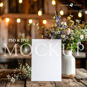 May include: A mockup image featuring a blank white card, a rustic vase filled with wildflowers, and a wooden table. The scene is illuminated by soft, warm lights, creating a cozy and inviting atmosphere. The text "PSD & JPG MOCKUP" is visible.