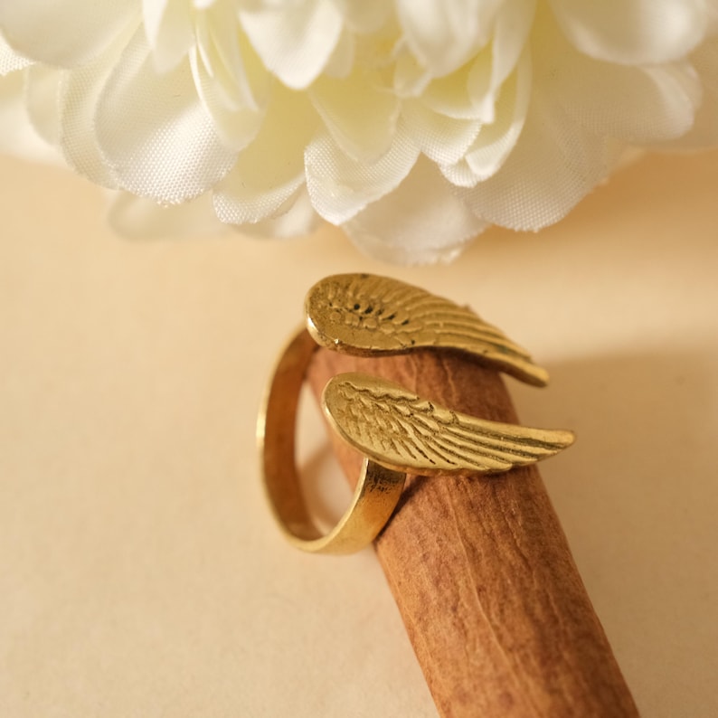 May include: A gold-colored ring with wing-shaped accents. The ring is set against a light brown background, with a white flower in the upper left corner. The wings are detailed with feather-like patterns.