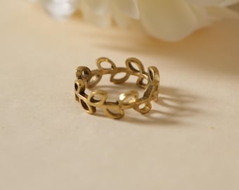 May include: A gold-colored ring with a leaf design. The ring is circular, with a series of connected leaf shapes forming the band. The ring is set against a soft, neutral background, with a blurred white flower in the upper left corner.