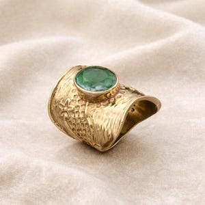 Peut inclure: Une bague dorée ornée d'une pierre précieuse ovale vert émeraude. La bague a un large anneau avec des détails floraux et texturés. La pierre est sertie dans une lunette surélevée, et le design est orné et détaillé.