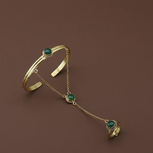 Op de afbeelding: Een goudkleurige handkettingarmband met groene edelstenen. De armband heeft een manchetontwerp dat door een fijne ketting met een ring is verbonden. De ring en armband hebben elk een groene edelsteen, met een extra edelsteen op de ketting.