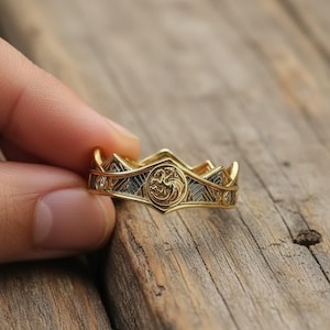 May include: A gold ring with a crown design. The ring features a central emblem with a dragon motif, surrounded by geometric patterns and small, clear stones. The ring is held against a wooden surface.