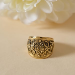 Può includere: Un anello largo color oro con un motivo a volute ornato. L'anello ha un design a fascia con dettagli a volute e piccoli accenti circolari. È posto su uno sfondo morbido e neutro con un fiore bianco sfocato nella parte superiore.
