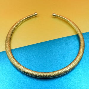 Puede incluir: Un collar dorado con un patrón texturizado en forma de escamas. El diseño abierto presenta pequeños terminales redondeados. El collar se muestra sobre un fondo amarillo y azul.