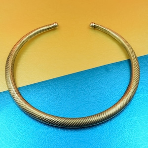 May include: A gold-coloured, open-ended collar necklace. The necklace features a textured, ridged design along its length. The ends are capped with small, rounded gold beads. The background is split yellow and blue.