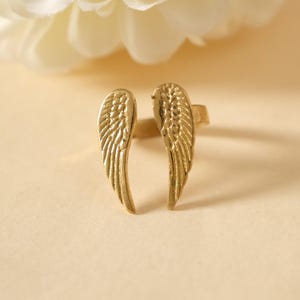 May include: A gold-colored ring featuring a design of two stylized wings. The wings are detailed with feather-like textures, and the ring is set against a soft, neutral background. The ring is a piece of jewelry.
