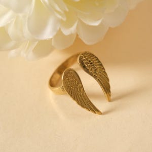 May include: A gold-colored ring with an open design featuring two detailed wing shapes. The ring is set against a soft, neutral background with a white flower in the upper left corner. The wings are approximately 1 inch in length.