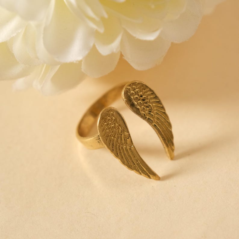 May include: A gold-colored ring with an open design featuring two detailed wing shapes. The ring is set against a soft, neutral background with a white flower in the upper left corner. The wings are approximately 1 inch in length.