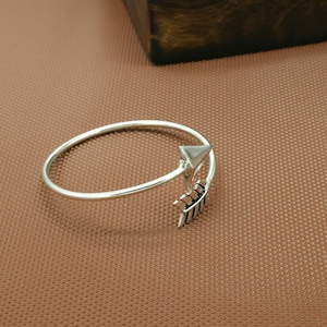 May include: A silver arrow bracelet. The bracelet has a simple band with an arrow design at the end. The arrow has a triangular head and detailed fletching. The bracelet is shown on a brown surface.
