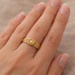 May include: A gold-colored ring with an intricate filigree design. The ring features a central circular element with a textured pattern. The band has an openwork design. The ring is on a person's finger.
