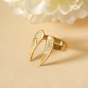 May include: A gold-colored ring with an open design featuring two wing-shaped elements. The wings have detailed feather patterns. The ring is set against a neutral background with a white flower in the upper corner.