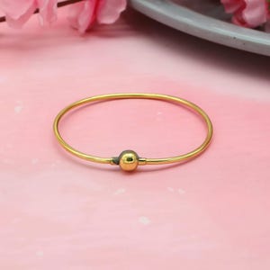 Op de afbeelding: Een goudkleurige armband met een kleine, ronde, goudkleurige kraal in het midden. De armband is eenvoudig en elegant, met een gladde, gepolijste afwerking. De armband is een sieraad.
