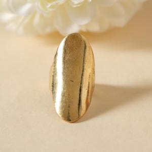 Op de afbeelding: Een goudkleurige ring met een langwerpig ovaal ontwerp. De ring heeft een glad, gepolijst oppervlak met kleine kleurvariaties, wat wijst op een vintage of handgemaakte uitstraling. De ring is een opvallend sieraad.
